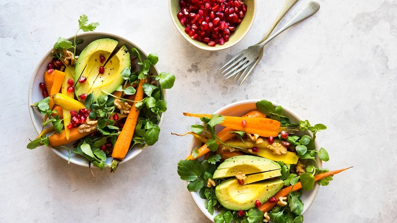 A colorful salad featuring sliced avocado, orange carrots, pomegranate seeds, walnuts, and fresh greens, served in two bowls with a small bowl of pomegranate seeds on the side.