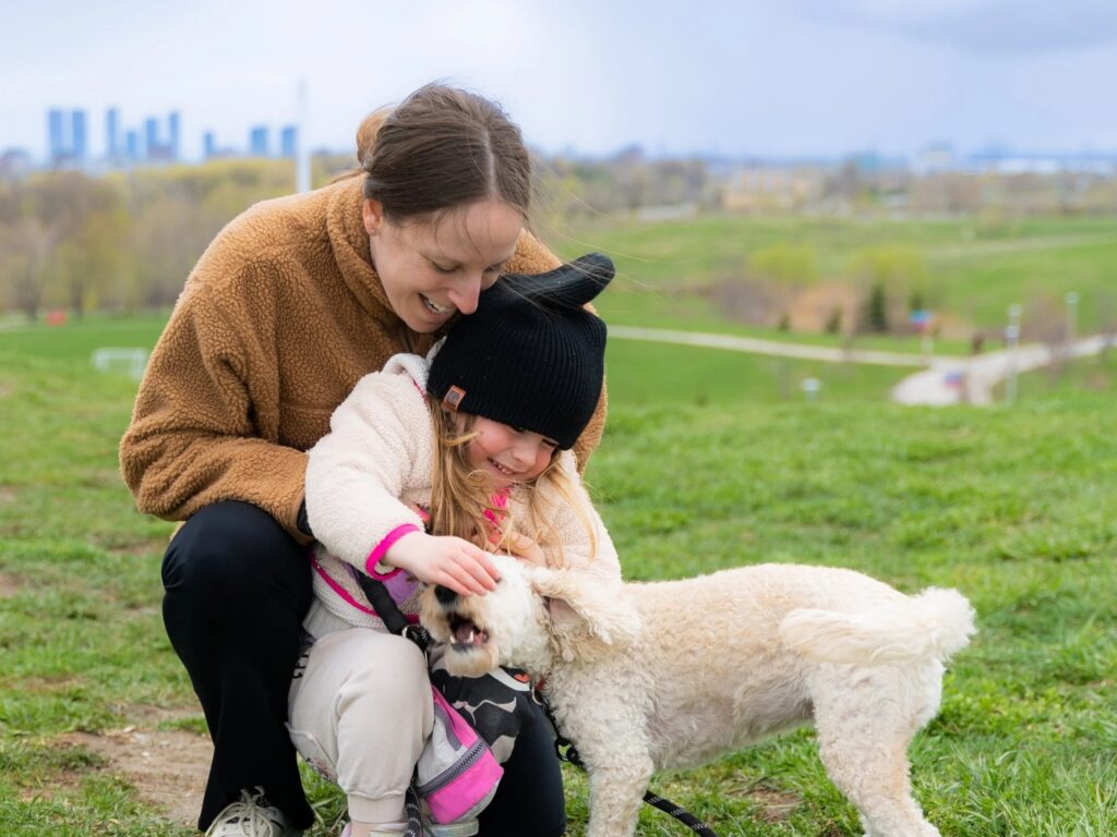 A mother and her daughter play with a dog in a grassy park, with a city skyline visible in the background.