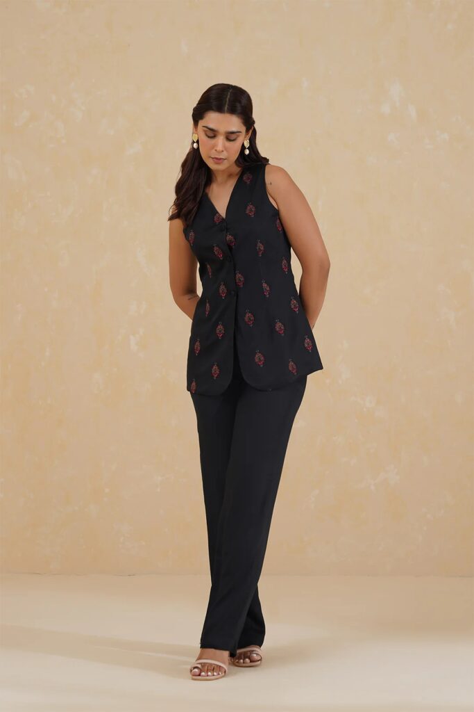A woman poses in a stylish sleeveless black top with red embroidery, paired with black pants and sandals, against a beige textured background.