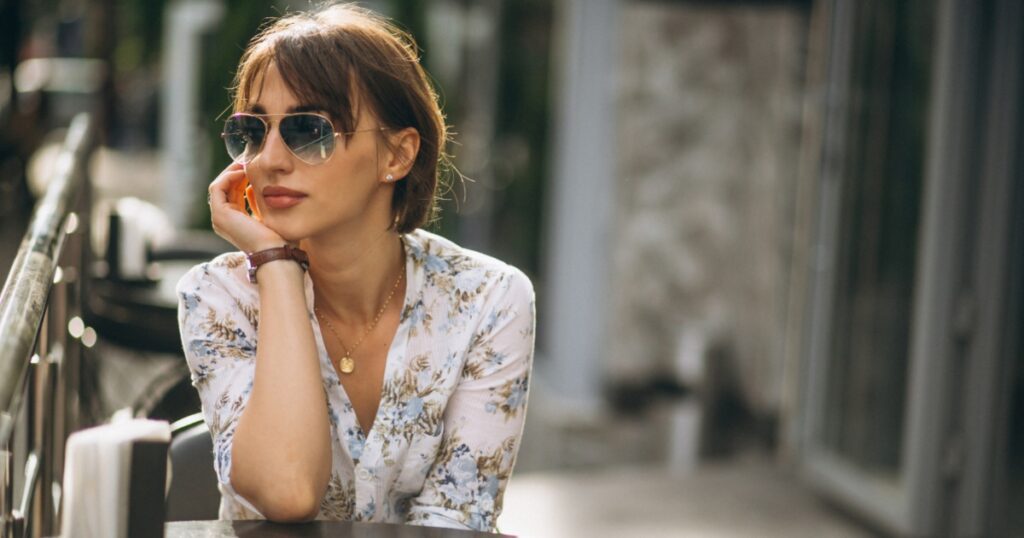 A young woman wearing sunglasses and a floral shirt, resting her chin on her hand while seated at an outdoor cafe.