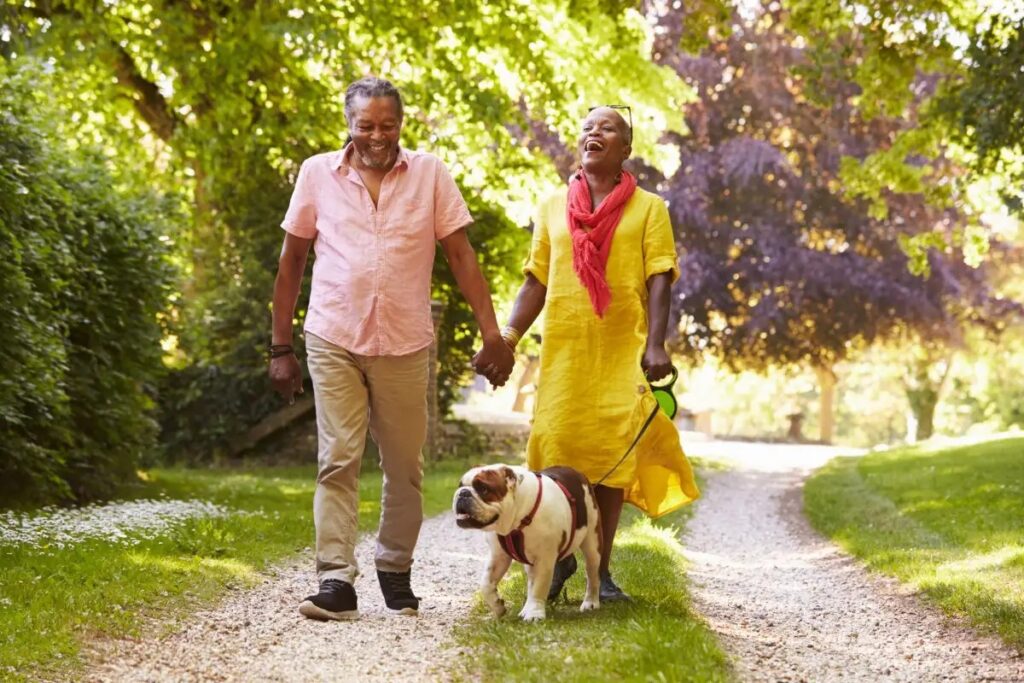 A happy couple walks hand in hand along a gravel path, accompanied by a playful bulldog, surrounded by lush greenery and vibrant trees.