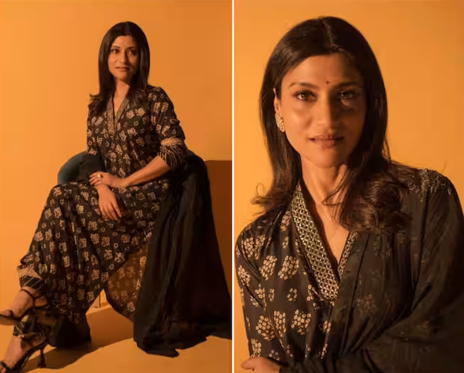 A woman in a stylish black outfit adorned with intricate patterns, sitting gracefully against a warm yellow backdrop, showcasing jewelry and a confident expression.