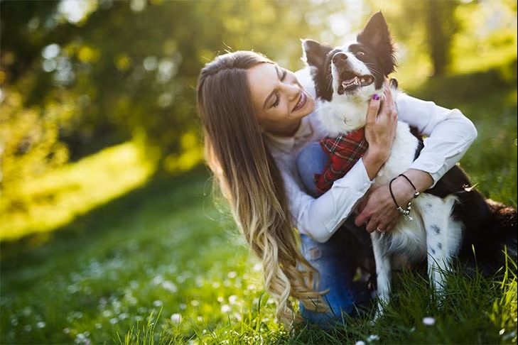 A woman embraces her dog in a sunlit grassy field, sharing a happy connection.