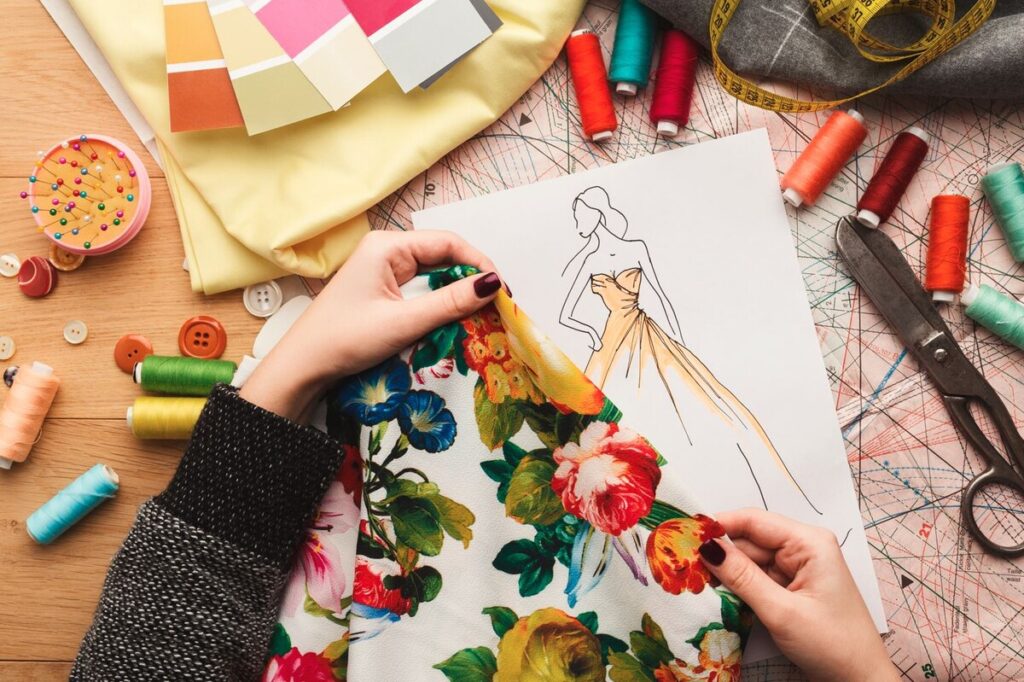 A person's hands exploring a floral fabric while sketching a dress design, surrounded by sewing supplies like threads, buttons, and patterns.