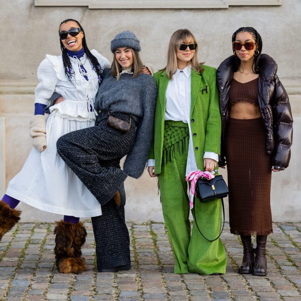 Four women pose joyfully in a range of trendy outfits, showcasing diverse styles against a cobblestone background.